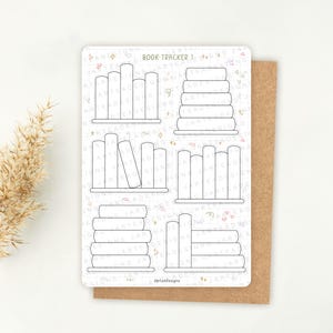 May include: A white sticker with the text "BOOK TRACKER 1" and illustrations of stacked books and bookshelves. The sticker is on a brown card, with a beige background and a dried plant in the corner.