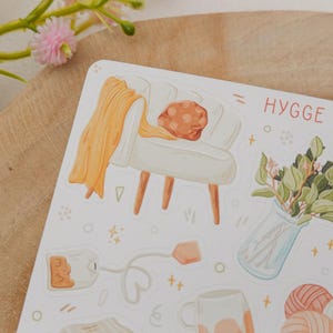Op de afbeelding: Een stickervel met illustraties van gezellige items. Het bevat een witte fauteuil met een oranje deken, een vaas met groen, een theezakje, een mok en garen met breinaalden. Het woord "HYGGE" is in het rood geschreven.
