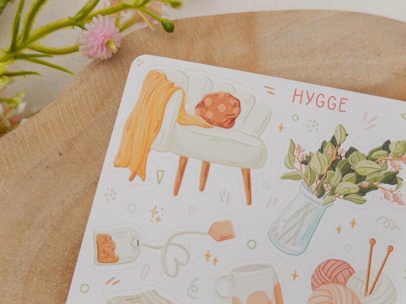 Op de afbeelding: Een stickervel met illustraties van gezellige items. Het bevat een witte fauteuil met een oranje deken, een vaas met groen, een theezakje, een mok en garen met breinaalden. Het woord "HYGGE" is in het rood geschreven.