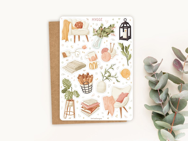 Op de afbeelding: Een stickerblad met een gezellig thema, met illustraties van een stoel, deken, boeken, planten en andere items. Het woord "HYGGE" staat bovenaan. Het stickerblad ligt op een bruine kaart.
