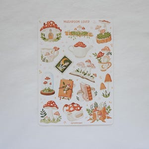 May include: A sticker sheet titled MUSHROOM LOVER featuring various illustrations of mushrooms, including a mushroom house, a teapot, mugs, books, and a glass dome. The designs are rendered in earthy tones of red, brown, green, and cream on a white background.
