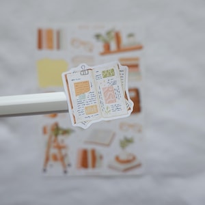 May include: A die-cut sticker featuring an open journal with handwritten notes, a binder clip, and decorative paper scraps in shades of beige, brown, and green. The sticker is held by a white pen, with a full sticker sheet visible in the background.