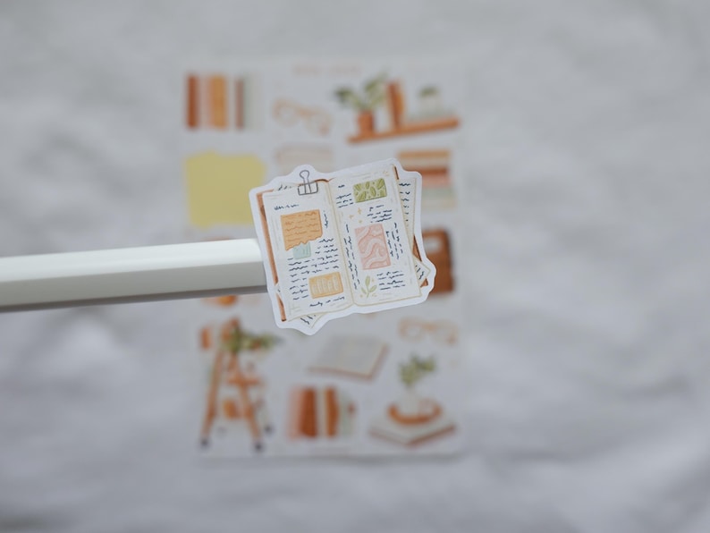 May include: A die-cut sticker featuring an open journal with handwritten notes, a binder clip, and decorative paper scraps in shades of beige, brown, and green. The sticker is held by a white pen, with a full sticker sheet visible in the background.