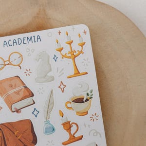 May include: A sticker sheet titled DARK ACADEMIA featuring illustrations of gold candelabras, a brown book, a white bust, round glasses, a steaming cup of coffee, a quill in an inkwell, a brown satchel, and white flowers with small star accents.