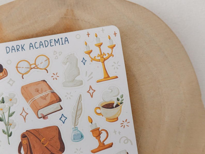 May include: A sticker sheet titled DARK ACADEMIA featuring illustrations of gold candelabras, a brown book, a white bust, round glasses, a steaming cup of coffee, a quill in an inkwell, a brown satchel, and white flowers with small star accents.