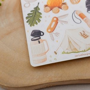 May include: A sticker sheet featuring camping illustrations including a beige thermos with a black cup, a white tent, an orange multi-tool, a green oak leaf, a campfire, binoculars, and a flashlight. The text JarteaDesigns is printed on the sheet.