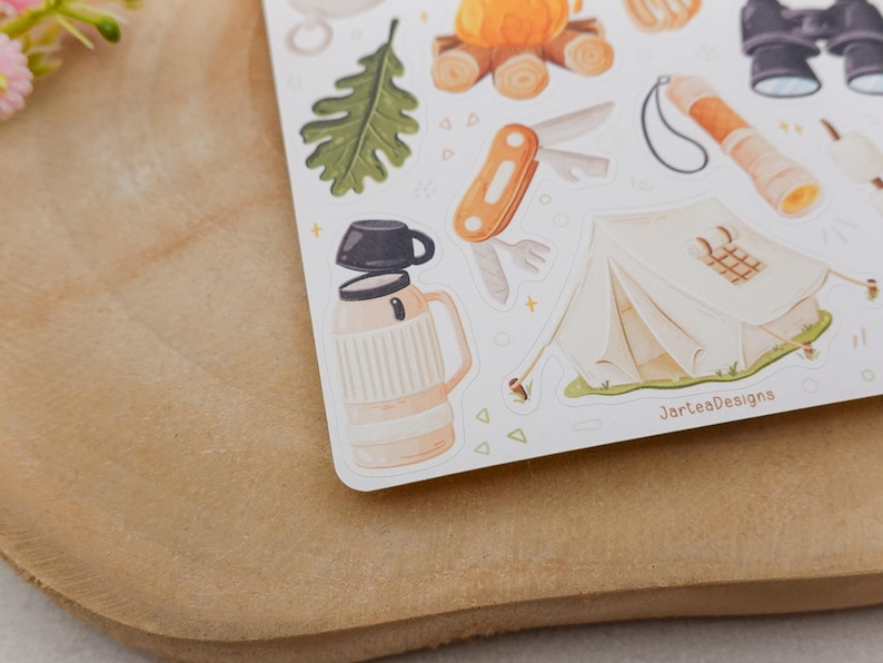 May include: A sticker sheet featuring camping illustrations including a beige thermos with a black cup, a white tent, an orange multi-tool, a green oak leaf, a campfire, binoculars, and a flashlight. The text JarteaDesigns is printed on the sheet.