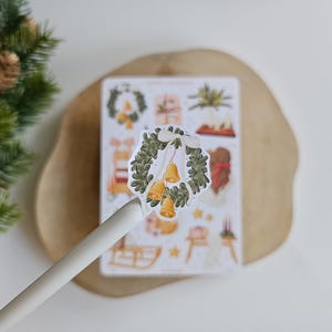 May include: A die-cut sticker featuring a green evergreen wreath adorned with a white ribbon and three golden bells. The sticker is held up by a white pen, positioned in front of a larger sticker sheet containing various holiday-themed illustrations.