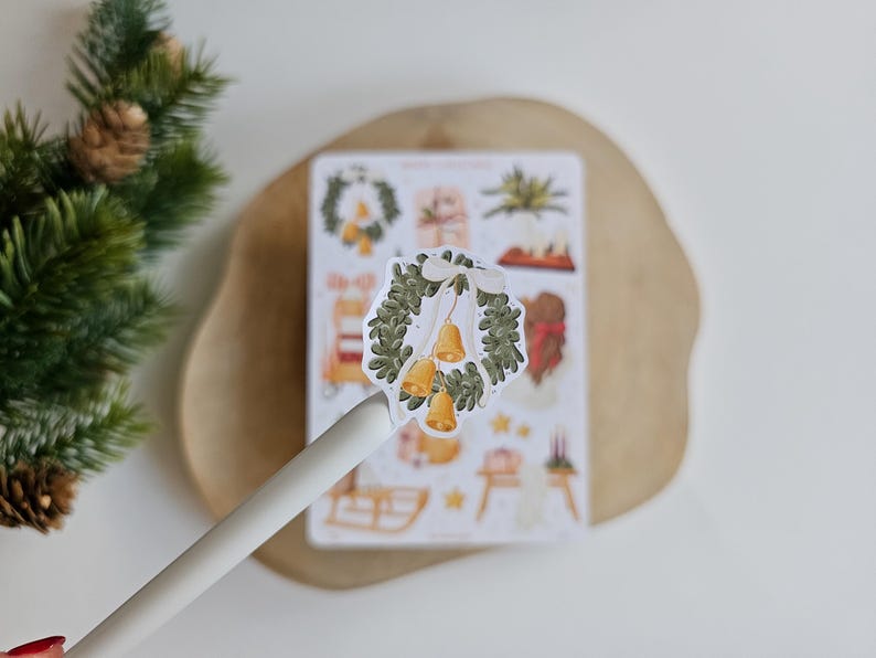 May include: A die-cut sticker featuring a green evergreen wreath adorned with a white ribbon and three golden bells. The sticker is held up by a white pen, positioned in front of a larger sticker sheet containing various holiday-themed illustrations.