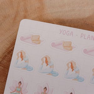 May include: A sheet of stickers with a yoga theme. The stickers feature illustrations of yoga poses, yoga mats, and blocks. The text "YOGA - PLANNER" is printed in a light purple color. The background is a light pink grid.