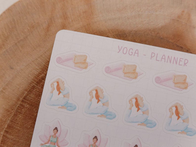 May include: A sheet of stickers with a yoga theme. The stickers feature illustrations of yoga poses, yoga mats, and blocks. The text "YOGA - PLANNER" is printed in a light purple color. The background is a light pink grid.