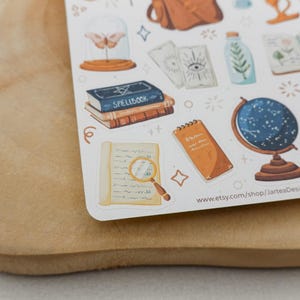 May include: A sticker sheet featuring various illustrations including a blue spellbook, a magnifying glass over a document, a celestial globe, tarot cards, a moth in a glass dome, and a potted plant, with the text www.etsy.com/shop/JarteaDesigns.