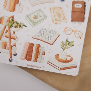 May include: A sticker sheet featuring various illustrations including a rolling cart with books and plants, an open book, a brown leather journal, light-colored glasses, and stacks of books with a small potted plant. The text JarteaDesigns is visible.