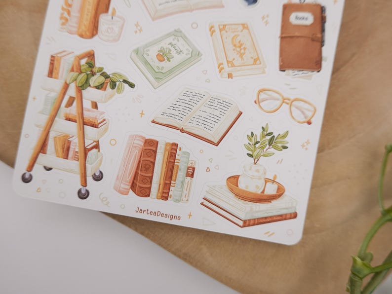 May include: A sticker sheet featuring various illustrations including a rolling cart with books and plants, an open book, a brown leather journal, light-colored glasses, and stacks of books with a small potted plant. The text JarteaDesigns is visible.