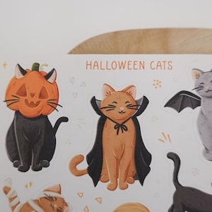 Pu&ograve; includere: Un foglio di adesivi intitolato HALLOWEEN CATS con illustrazioni di gatti in costume. Un gatto indossa una testa di zucca, un altro un mantello nero e un terzo ha ali da pipistrello. Sono presenti anche un gatto mummia e uno con un cappello viola.