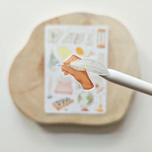 Puede incluir: Una pegatina troquelada que muestra una mano de color bronceado sosteniendo una pluma blanca. La pegatina est&aacute; colocada en la punta de un l&aacute;piz &oacute;ptico blanco, con una hoja de pegatinas rectangular que contiene varias ilustraciones peque&ntilde;as y coloridas al fondo.