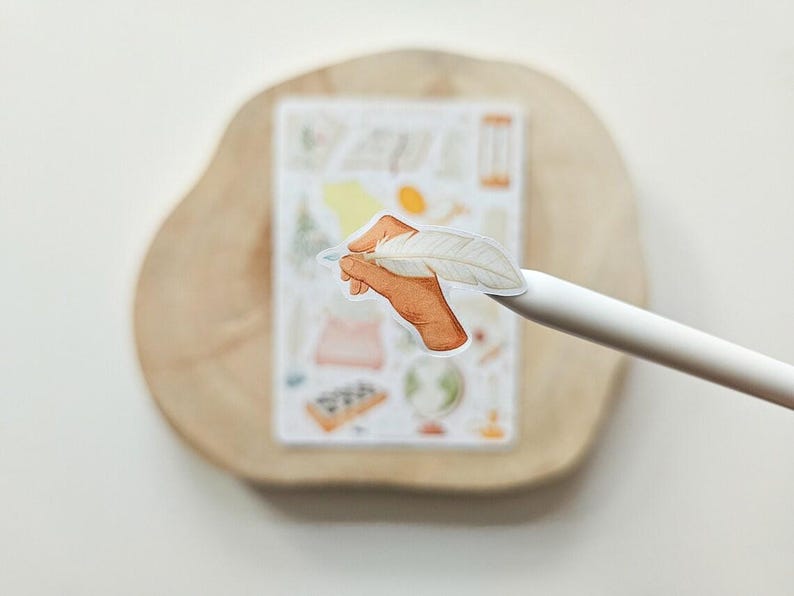 Puede incluir: Una pegatina troquelada que muestra una mano de color bronceado sosteniendo una pluma blanca. La pegatina est&aacute; colocada en la punta de un l&aacute;piz &oacute;ptico blanco, con una hoja de pegatinas rectangular que contiene varias ilustraciones peque&ntilde;as y coloridas al fondo.