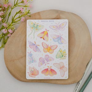 May include: A rectangular sticker sheet titled MAGICAL MOTHS featuring various pastel-colored moth illustrations in shades of pink, yellow, blue, and green. The moths are arranged in a grid pattern with small star accents on a white background.