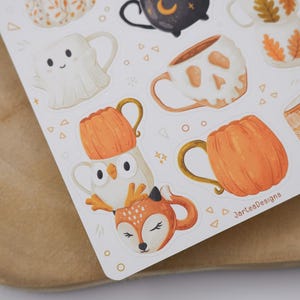 May include: A sticker sheet with illustrations of mugs in autumnal and Halloween designs. The mugs feature a ghost, a skull, pumpkins, and woodland animals. The colour scheme is mainly orange, white, and brown.