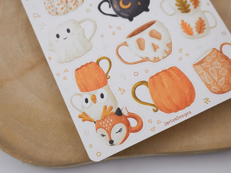 May include: A sticker sheet with illustrations of mugs in autumnal and Halloween designs. The mugs feature a ghost, a skull, pumpkins, and woodland animals. The colour scheme is mainly orange, white, and brown.