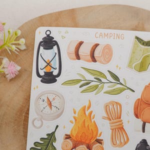May include: A sticker sheet featuring camping-themed illustrations including a black lantern, a rolled sleeping bag, a green leaf, a compass, a campfire, a coil of rope, a brown backpack, and a map, with the word CAMPING printed at the top.