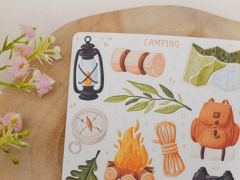May include: A sticker sheet featuring camping-themed illustrations including a black lantern, a rolled sleeping bag, a green leaf, a compass, a campfire, a coil of rope, a brown backpack, and a map, with the word CAMPING printed at the top.