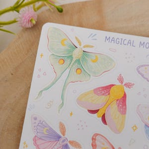 May include: A sticker sheet titled MAGICAL MOTHS featuring watercolor illustrations of various moths in pastel shades of green, pink, yellow, and purple. Each moth is decorated with small crescent moon and circular patterns on its wings.