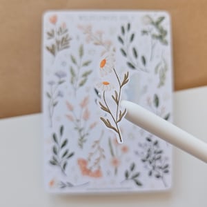 May include: A white sticker sheet titled WILDFLOWERS VOL. 2 featuring various botanical illustrations of green leaves, stems, and small flowers. A single daisy sticker is being lifted from the sheet by a white stylus, showing two white petals with yellow centers.