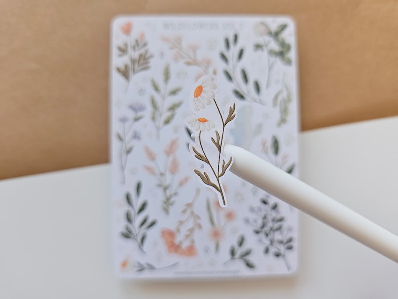 May include: A white sticker sheet titled WILDFLOWERS VOL. 2 featuring various botanical illustrations of green leaves, stems, and small flowers. A single daisy sticker is being lifted from the sheet by a white stylus, showing two white petals with yellow centers.