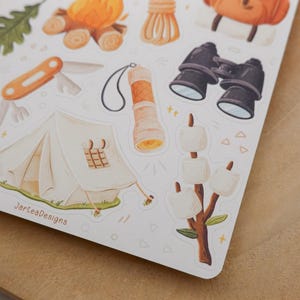 May include: A sticker sheet featuring camping-themed illustrations including a white tent, a campfire, a multi-tool, a flashlight, binoculars, and marshmallows on a stick. The text JarteaDesigns is printed in brown near the bottom left corner.