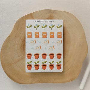 May include: A sticker sheet with a plant care planner design. The sheet features illustrations of watering cans, seed packets, potted plants, and sprouting plants. The text "PLANT CARE - PLANNER" is at the top. The sticker sheet is on a wooden surface.