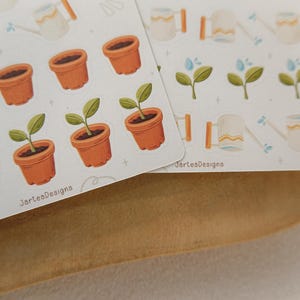 May include: Two sheets of stickers with gardening illustrations. The stickers depict terracotta pots with plants, watering cans, and sprouting plants. The text "JarteaDesigns" is printed on the bottom of the sheets.