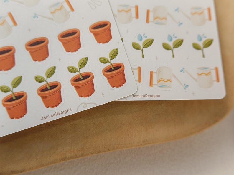 May include: Two sheets of stickers with gardening illustrations. The stickers depict terracotta pots with plants, watering cans, and sprouting plants. The text "JarteaDesigns" is printed on the bottom of the sheets.