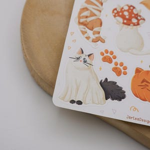 Pu&ograve; includere: Un foglio di adesivi con illustrazioni di un gatto bianco vestito da fantasma, una zucca con faccia da gatto, impronte di zampe arancioni e un fungo rosso a pois bianchi. Il testo JarteaDesigns &egrave; stampato nell'angolo in basso a destra.
