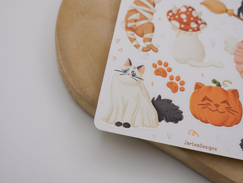 Pu&ograve; includere: Un foglio di adesivi con illustrazioni di un gatto bianco vestito da fantasma, una zucca con faccia da gatto, impronte di zampe arancioni e un fungo rosso a pois bianchi. Il testo JarteaDesigns &egrave; stampato nell'angolo in basso a destra.