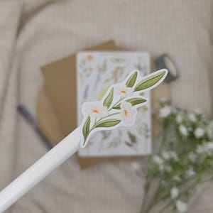 May include: A white pen featuring a die-cut sticker of a botanical illustration. The sticker displays three pale pink flowers with yellow centers and green leaves attached to a slender stem, rendered in a clean, flat graphic style on a white background.
