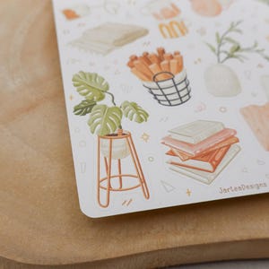 Op de afbeelding: Een stickervel met aquarel illustraties van woondecoratie-items. De ontwerpen omvatten een plant in pot, boeken, een gevlochten mand met stokken en een vaas met groen. Het kleurenpalet is zacht, met tinten oranje, groen en beige.
