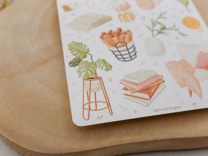 Op de afbeelding: Een stickervel met aquarel illustraties van woondecoratie-items. De ontwerpen omvatten een plant in pot, boeken, een gevlochten mand met stokken en een vaas met groen. Het kleurenpalet is zacht, met tinten oranje, groen en beige.