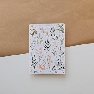 May include: A rectangular sticker sheet featuring various botanical illustrations of wildflowers and foliage in muted tones of green, pink, and white. The text WILDFLOWERS VOL. 2 is printed at the top, and small star and circle accents are scattered throughout.