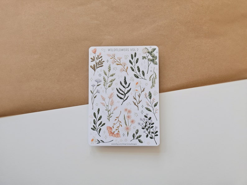 May include: A rectangular sticker sheet featuring various botanical illustrations of wildflowers and foliage in muted tones of green, pink, and white. The text WILDFLOWERS VOL. 2 is printed at the top, and small star and circle accents are scattered throughout.