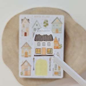 May include: A die-cut sticker of a two-story house with a dark brown roof covered in white snow, featuring yellow-lit windows, a central door, and string lights. Behind it is a sticker sheet labeled Winter Village with various house illustrations.