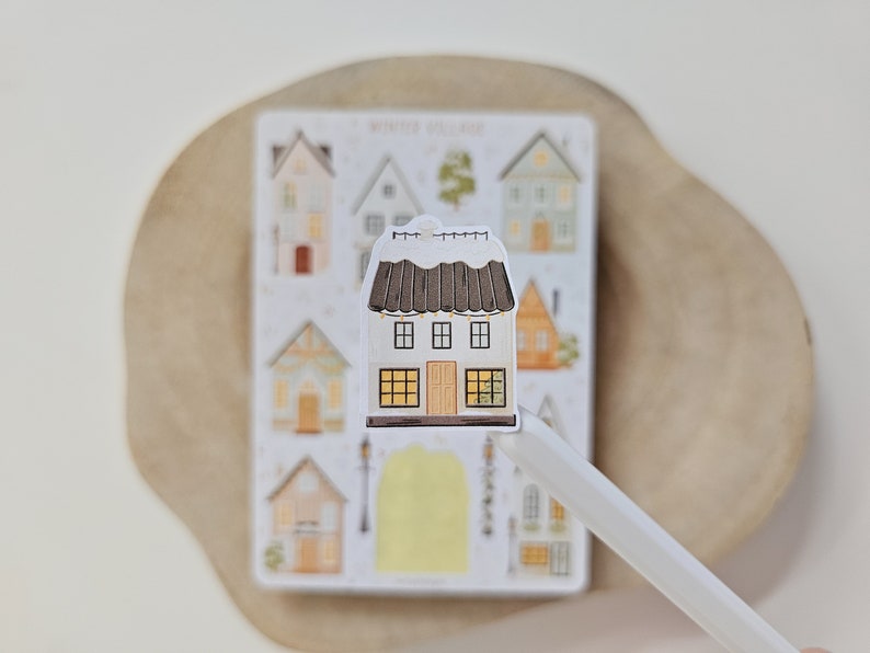 May include: A die-cut sticker of a two-story house with a dark brown roof covered in white snow, featuring yellow-lit windows, a central door, and string lights. Behind it is a sticker sheet labeled Winter Village with various house illustrations.