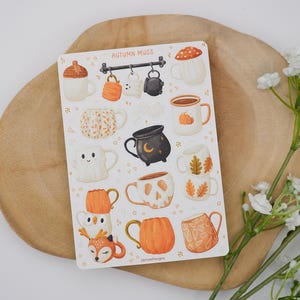 May include: A sticker sheet with illustrations of autumn-themed mugs. The mugs have designs such as pumpkins, acorns, and woodland creatures. The color scheme is warm, with orange, brown, and cream tones. The text "AUTUMN MUGS" is displayed at the top of the sheet.