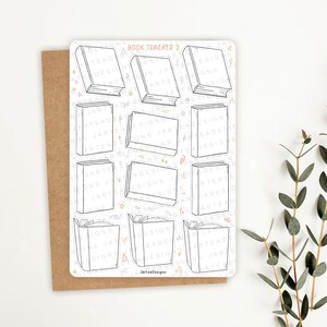 May include: A white sticker sheet with the text "Book Tracker 2" and outlines of books. The sticker sheet is on a brown card with a white background and a sprig of green leaves.
