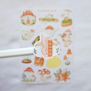 May include: A sticker featuring two mushrooms flanking a book titled Mycology, held on the tip of a white pencil. In the background, a sticker sheet displays various illustrations of mushrooms, teapots, and books in soft orange, green, and brown tones.