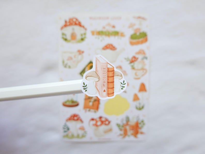 May include: A sticker featuring two mushrooms flanking a book titled Mycology, held on the tip of a white pencil. In the background, a sticker sheet displays various illustrations of mushrooms, teapots, and books in soft orange, green, and brown tones.