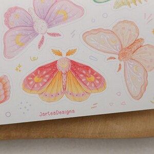 May include: A sticker sheet featuring multiple moth illustrations in pastel shades of pink, orange, and purple, each decorated with yellow crescent moon symbols. The text JarteaDesigns is printed in small pink letters at the bottom of the sheet.