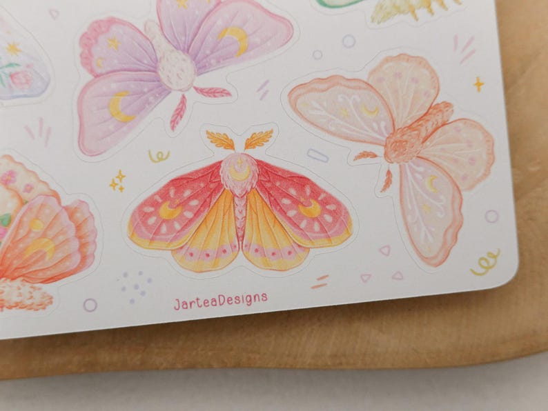 May include: A sticker sheet featuring multiple moth illustrations in pastel shades of pink, orange, and purple, each decorated with yellow crescent moon symbols. The text JarteaDesigns is printed in small pink letters at the bottom of the sheet.
