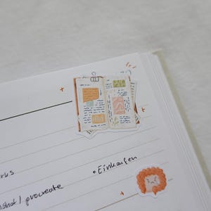 May include: A white notebook page featuring two stickers. One sticker depicts an open journal with handwritten notes, orange and green paper scraps, and a silver binder clip. The second sticker is a small, round, orange, ruffled character.