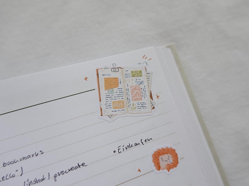 May include: A white notebook page featuring two stickers. One sticker depicts an open journal with handwritten notes, orange and green paper scraps, and a silver binder clip. The second sticker is a small, round, orange, ruffled character.